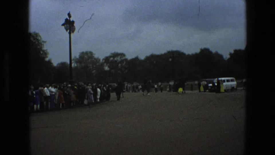 1969 England home-movie frame with onlookers beside a broad paved road, mounted police in the distance, trees, a lamppost, and cloudy sky.