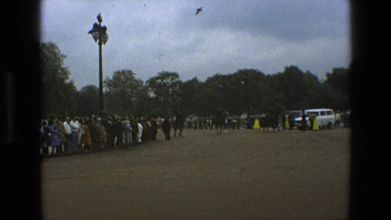 1969 England frame showing a crowd beside a paved park road, mounted police farther back, a white service vehicle, trees, and an ornamental lamppost.