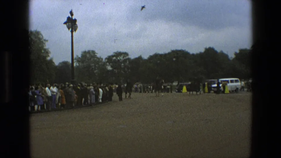 1969 England frame showing a crowd beside a paved park road, mounted police farther back, a white service vehicle, trees, and an ornamental lamppost.