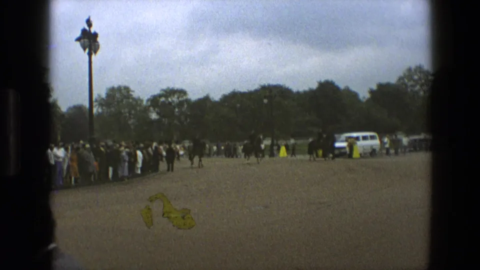 1969 England home-movie still with spectators at the edge of a park road, distant mounted police, a white vehicle, trees, and a tall lamppost.