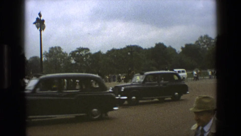 1969 England frame with black period cars crossing the foreground, onlookers and mounted police behind them, trees, a lamppost, and cloudy sky.