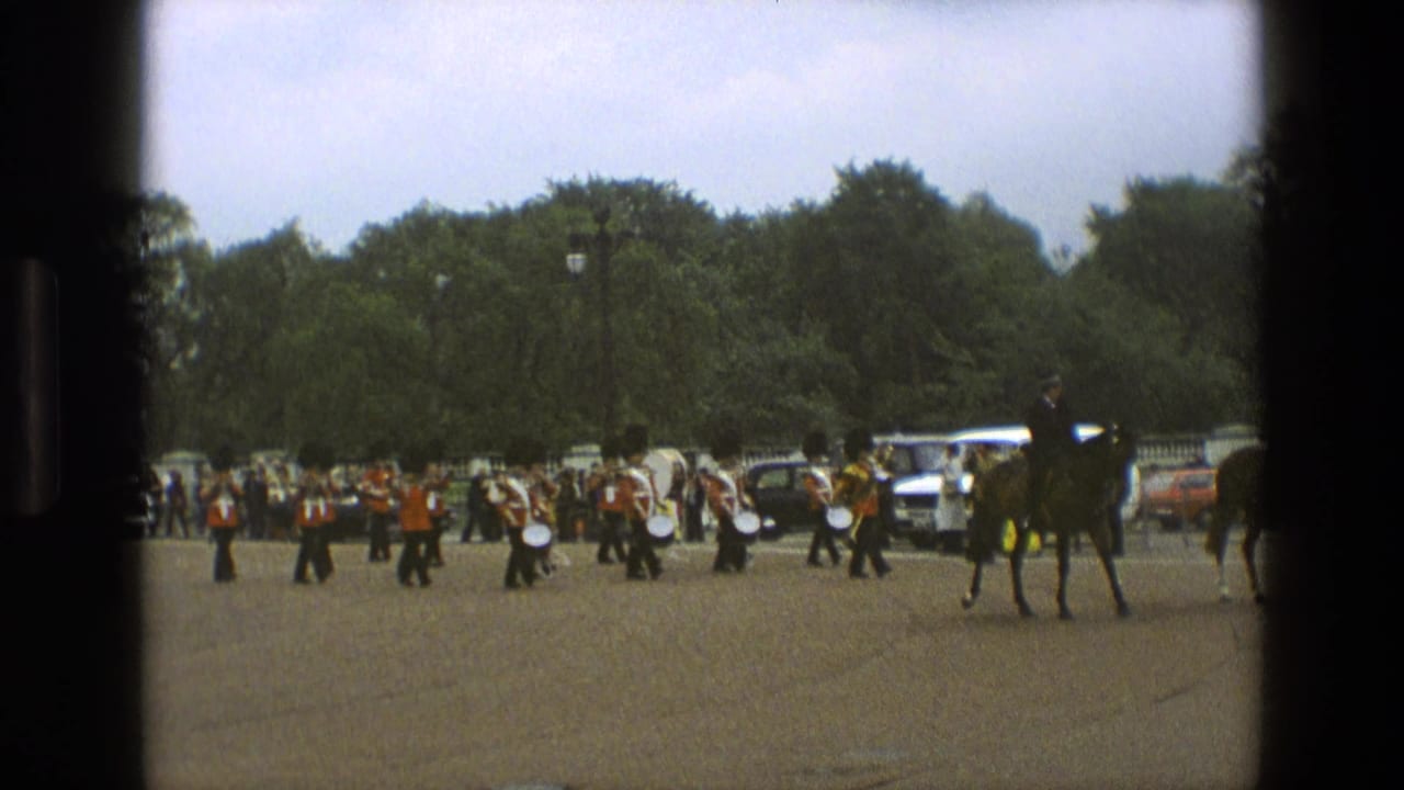 1969 England 8mm frame showing a mounted rider leading or passing a red-coated band on open paved grounds with spectators and cars in the background.