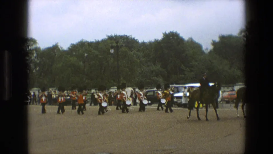 1969 England 8mm frame showing a mounted rider leading or passing a red-coated band on open paved grounds with spectators and cars in the background.