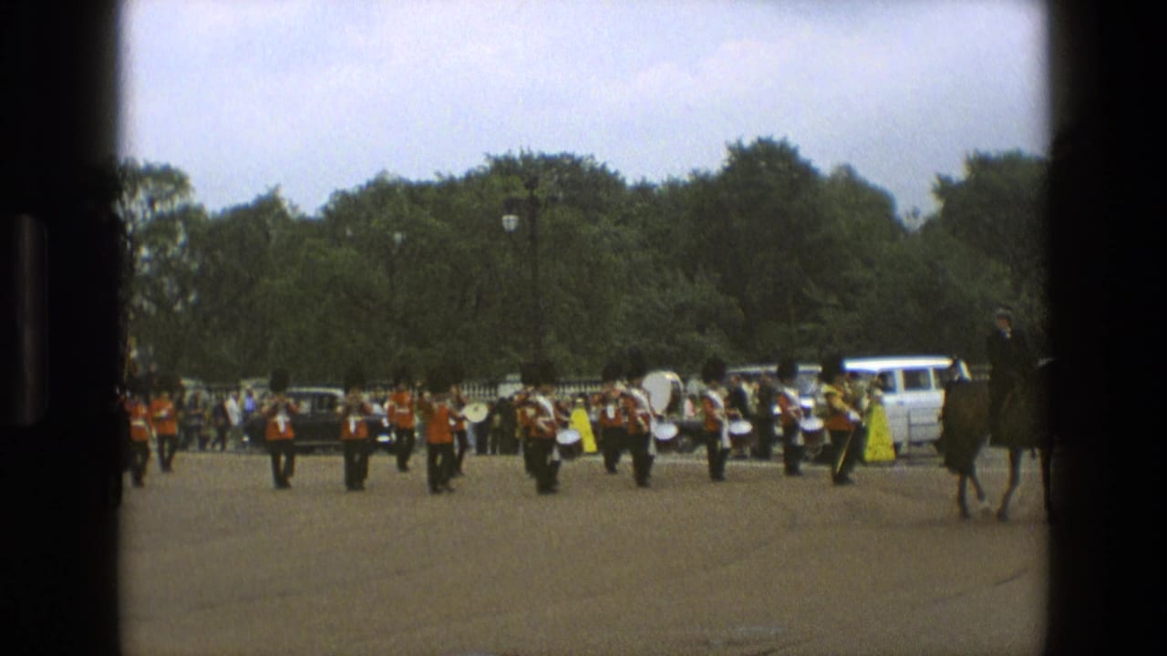 Red-coated marching band with drums moves across an open area, with mounted riders at the edges.