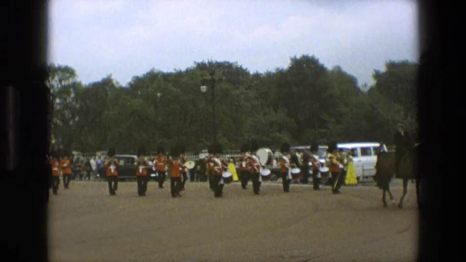 1969 England home-movie frame of a red-coated marching band with percussion moving across a broad paved space, watched by spectators near parked vehicles.