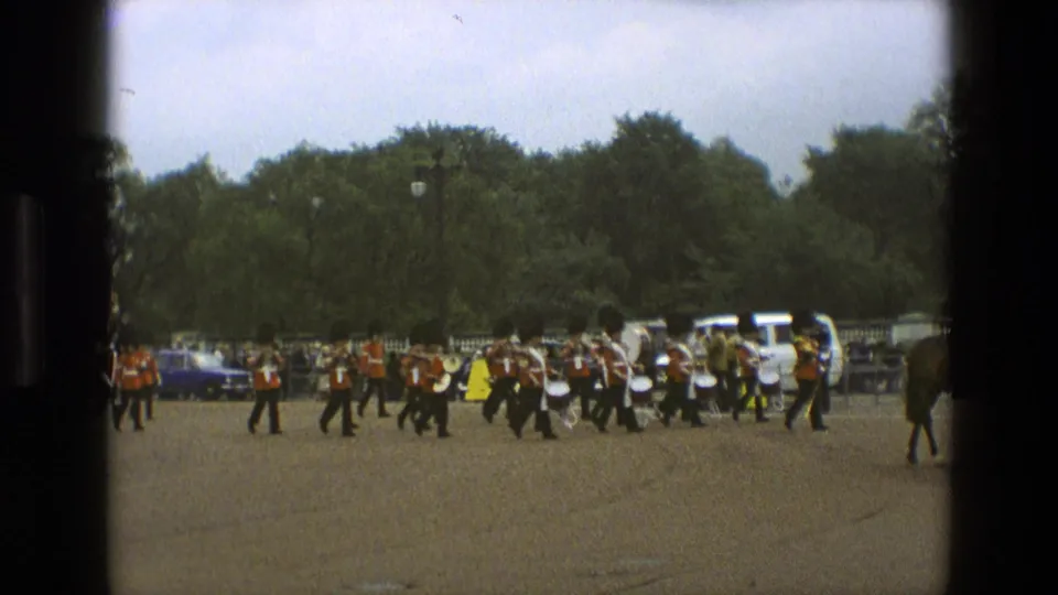 1969 England 8mm frame showing the band percussion section in red coats and dark trousers marching past a line of spectators and parked cars.