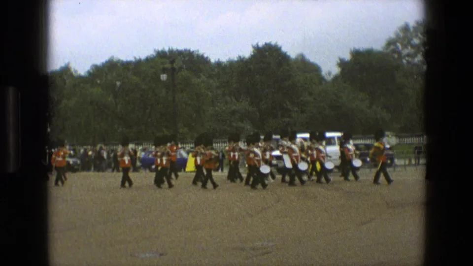 1969 England home-movie frame with red-coated band members marching left to right, drums visible, a horse at the edge and spectators behind the route.