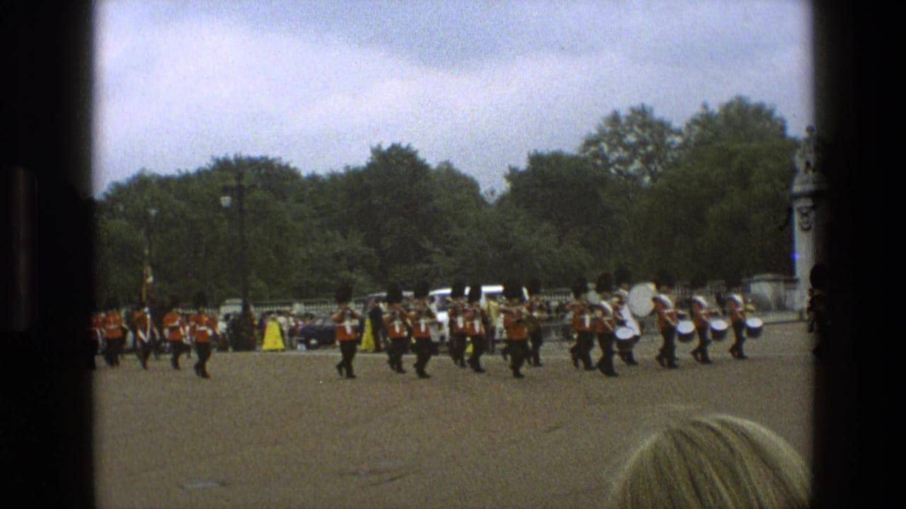 A formation of guards and drummers marches past trees, cars, and onlookers.