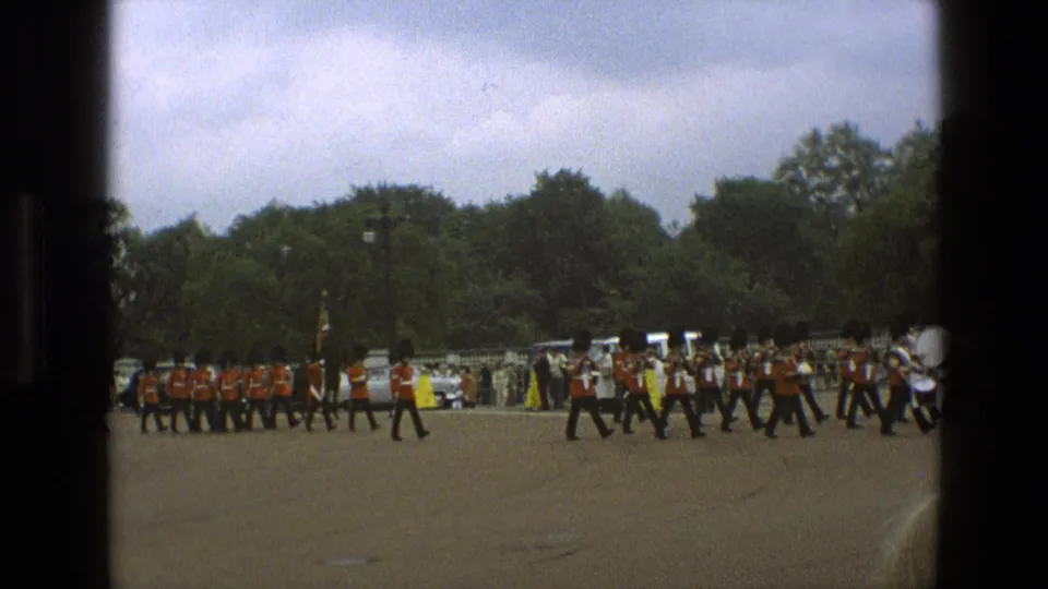1969 England home-movie frame with red-coated guards marching in rows across a public square, backed by trees, cars, and onlookers.