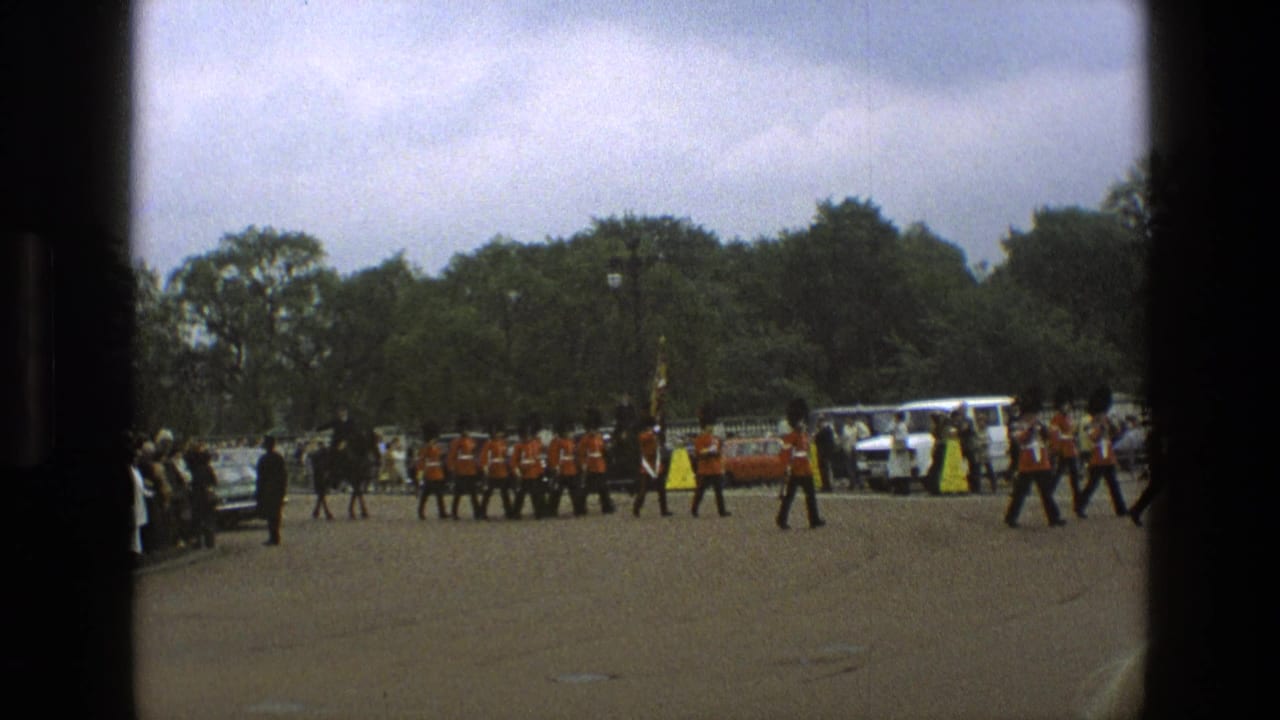 1969 England home-movie frame showing guards in red tunics marching in formation with spectators and cars nearby.