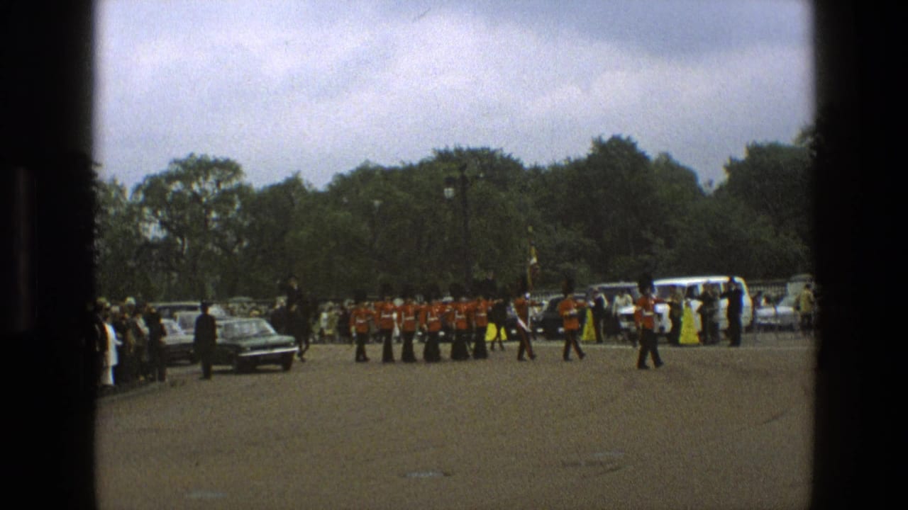 Marching guards pass a car and roadside spectators under an overcast sky.