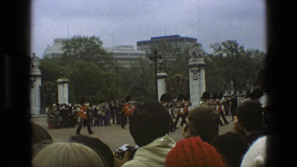 Visitor-shot 1969 England frame with crowd heads in the foreground, guards near palace gates, trees, and distant buildings.