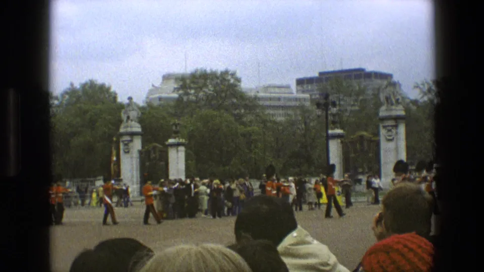 1969 England home-movie view of guards, spectators, ornate gates, trees, and foreground onlookers during a public ceremony.