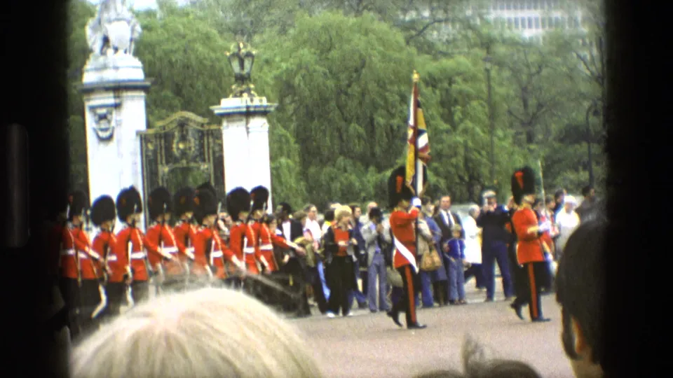 Closer 1969 England view of guards in red tunics near an ornate gate, with visitors and trees visible behind the line.