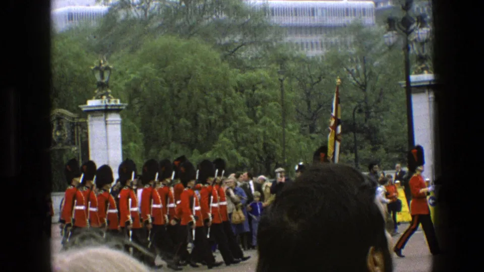 1969 England home-movie frame of guards in red tunics marching past spectators near gates and green trees.