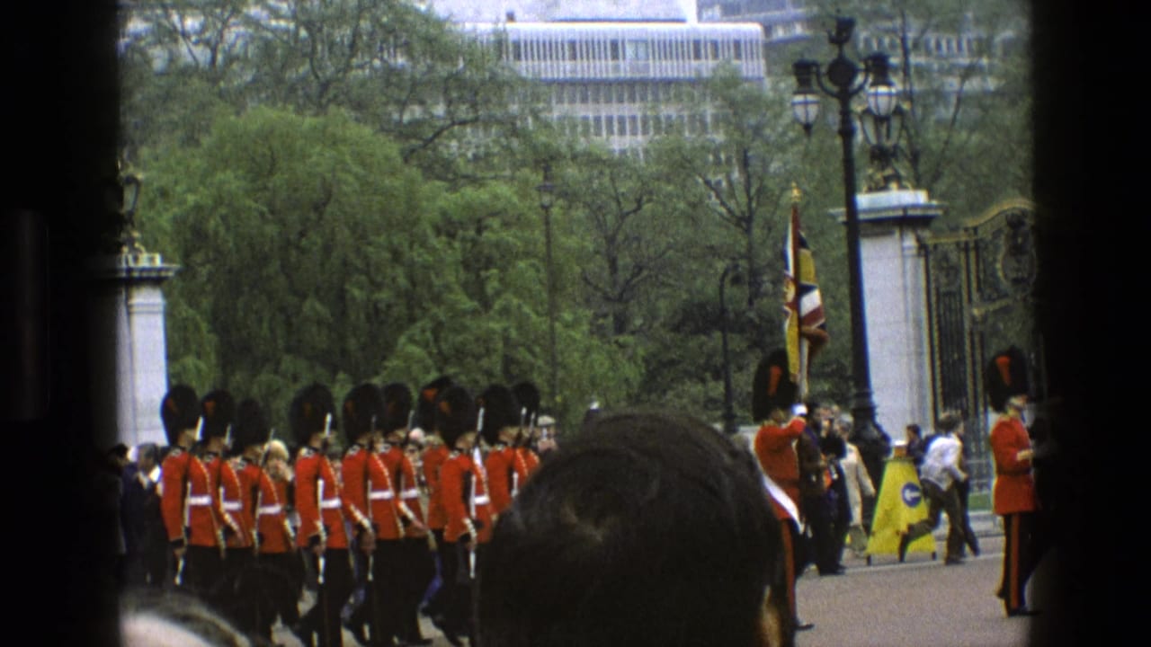 1969 England home-movie frame showing Queen's Guards marching in formation near ornate gates while spectators watch from the foreground.