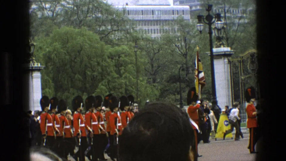 1969 England home-movie frame showing Queen's Guards marching in formation near ornate gates while spectators watch from the foreground.