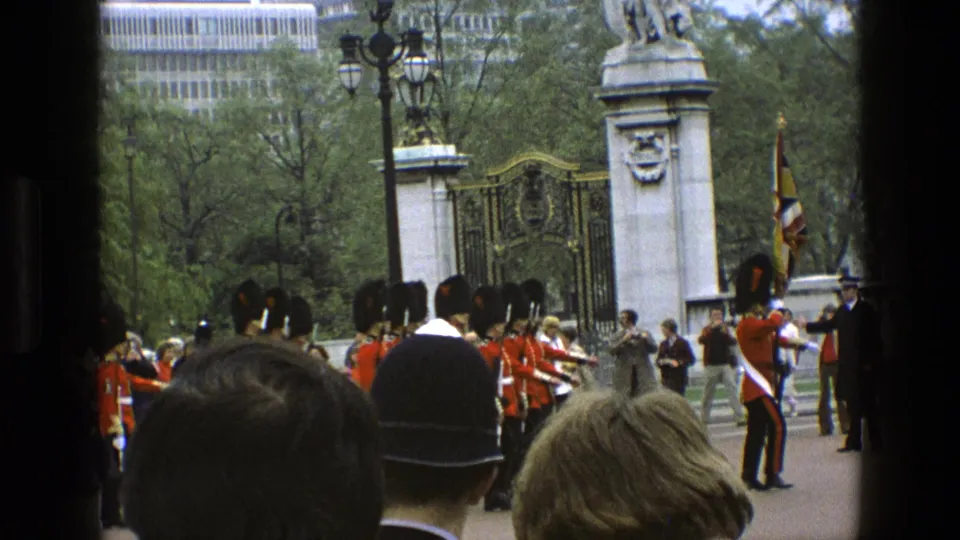1969 England home-movie frame showing a guard holding a flag near marching guards, foreground spectators, stone pillars, and park trees.