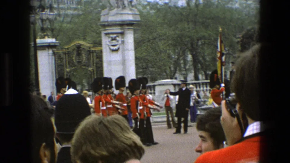1969 England home-movie frame showing guards in red tunics near ornate gates, with spectators in the foreground and trees behind.