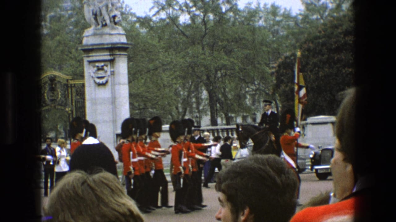 1969 England home-movie frame of guards in red tunics and bearskin-style hats marching near ornate gates as spectators watch.