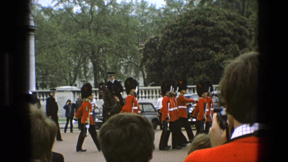 Visitor-shot 1969 England frame showing guards marching in formation beside spectators, parked cars, and formal gate architecture.