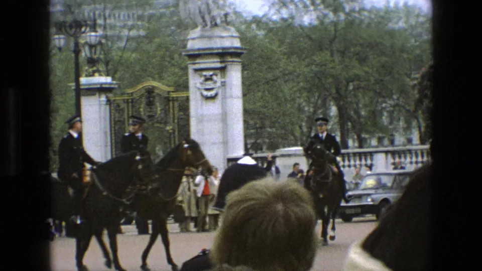 1969 England home-movie frame with mounted police riding past spectators and cars near ornate stone gate pillars.