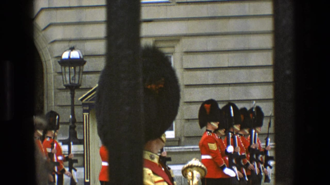 British Royal Guard members in red coats and bearskin hats stand in formation outside a grand stone building in England.