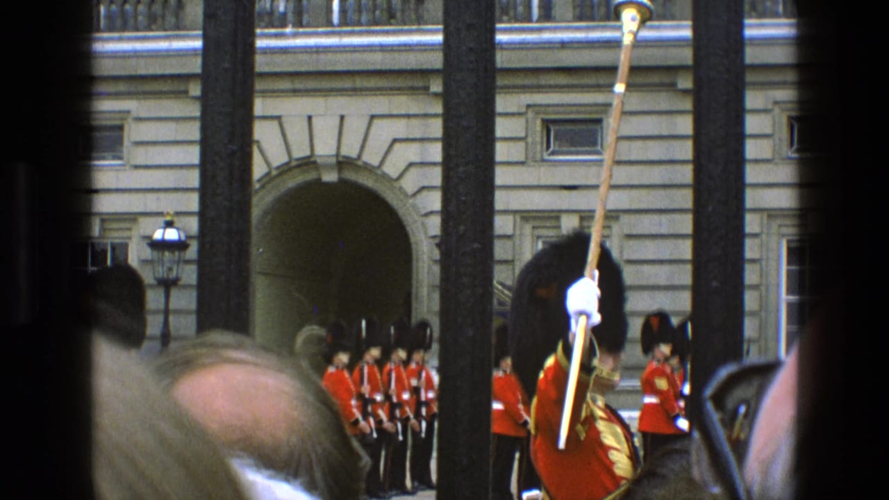 1969 London home-movie view of Queen's Guard at Buckingham Palace through iron railings, partly blocked by spectators.