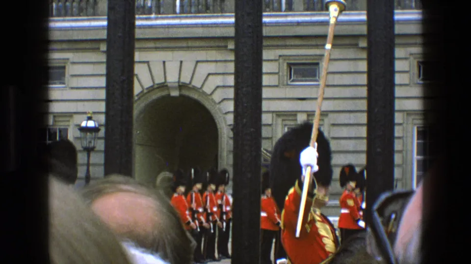 1969 London home-movie view of Queen's Guard at Buckingham Palace through iron railings, partly blocked by spectators.