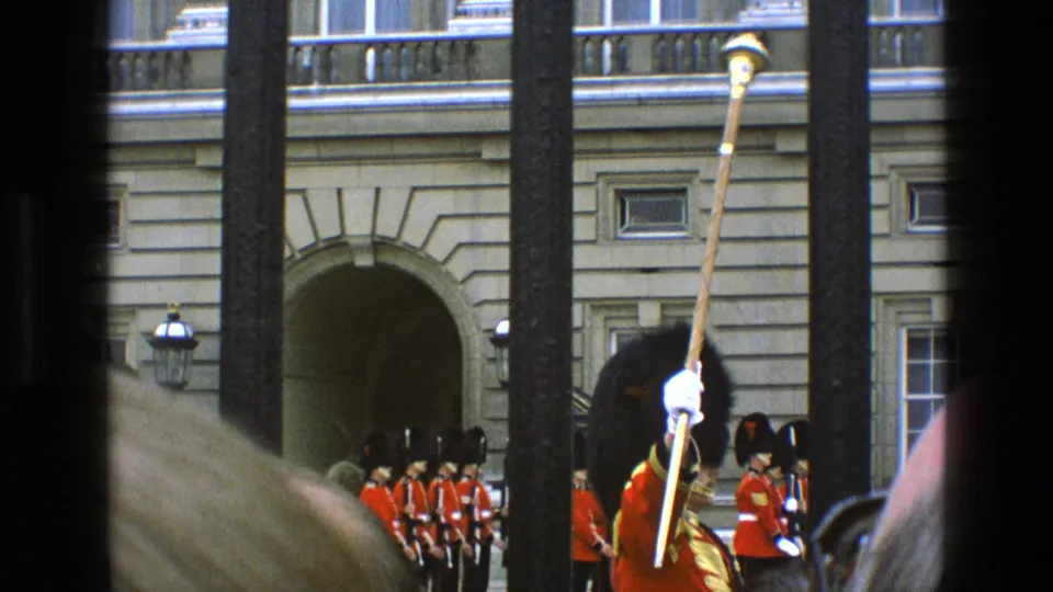 Visitor-shot 8mm frame of Buckingham Palace guards partly obscured by foreground spectators and iron bars.