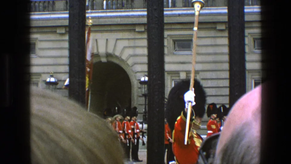 Buckingham Palace guard scene with red uniforms, iron railings, spectator heads, and a brief flag in 1969 footage.