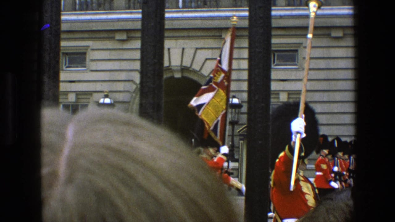 1969 England home-movie frame showing ceremonial guards, a flag, spectators, and vertical railings before a formal stone building.