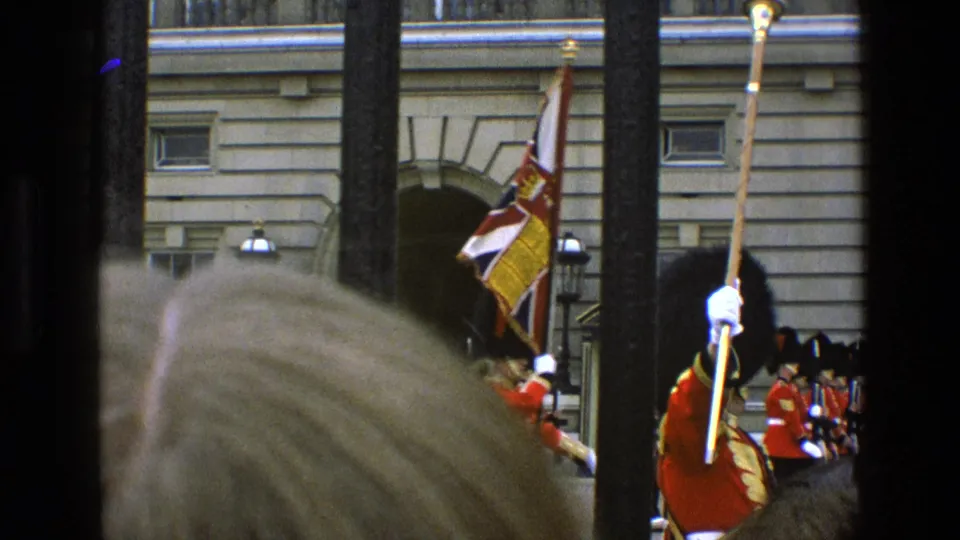 1969 England home-movie frame showing ceremonial guards, a flag, spectators, and vertical railings before a formal stone building.