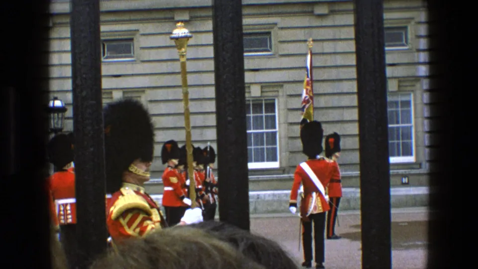 1969 England frame of red-coated ceremonial guards and a flag near a stone palace-like facade, viewed past dark railings.