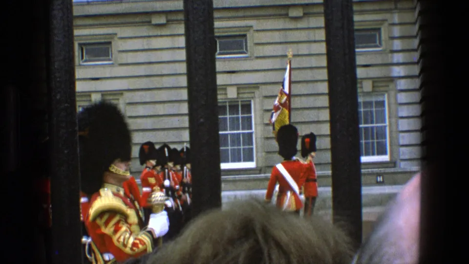 1969 England home-movie still with a gold-trimmed ceremonial guard in the foreground and red-coated guards beyond railings.
