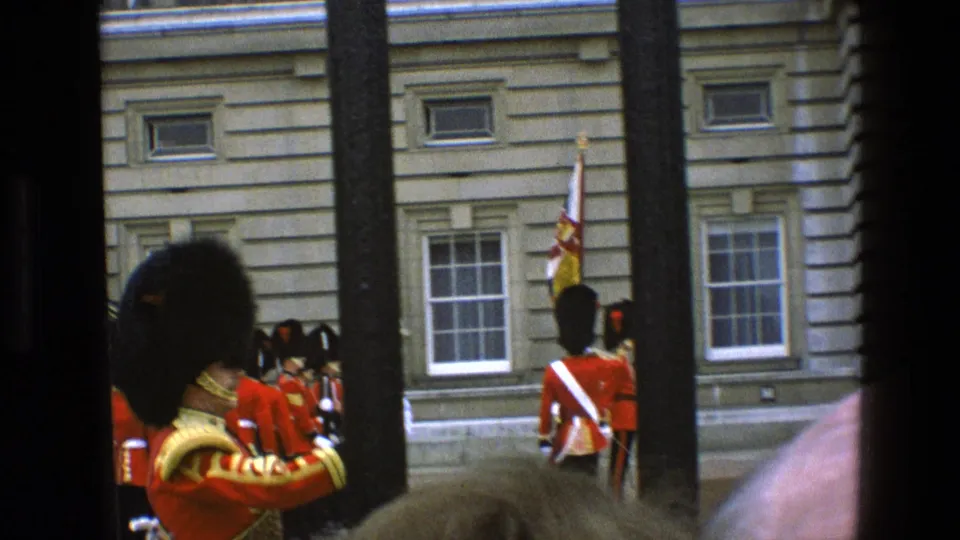 1969 England frame showing a red-uniformed guard from behind, other ceremonial guards, a flag, and palace-area railings.