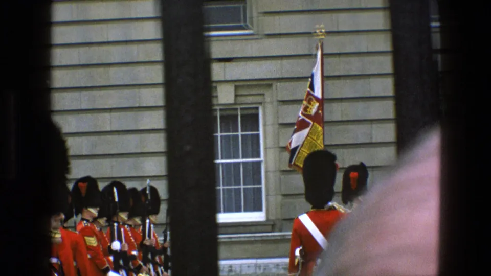 1969 England 8mm frame with red-coated guards, a royal flag and stone facade, partially blocked by bars and foreground blur.