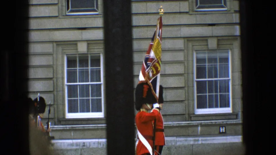 1969 England frame showing a red-coated ceremonial guard holding a royal flag outside a stone palace facade behind black bars.