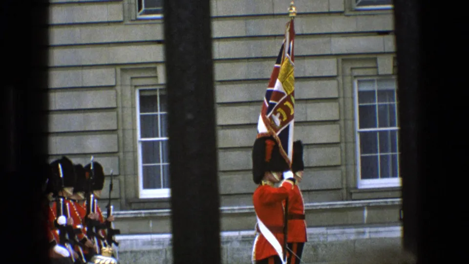 1969 England footage shows Grenadier Guards in ceremonial red tunics and bearskin hats near Buckingham Palace, partly obscured by dark bars.