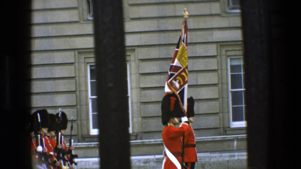 At Buckingham Palace in 1969, a ceremonial guard holds a Union Jack standard as other red-uniformed guards stand in formation behind bars.