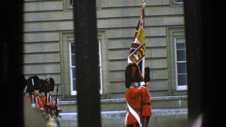 Handheld 8mm footage shows a Buckingham Palace guard with a standard and nearby soldiers in red ceremonial dress, seen through vertical bars.