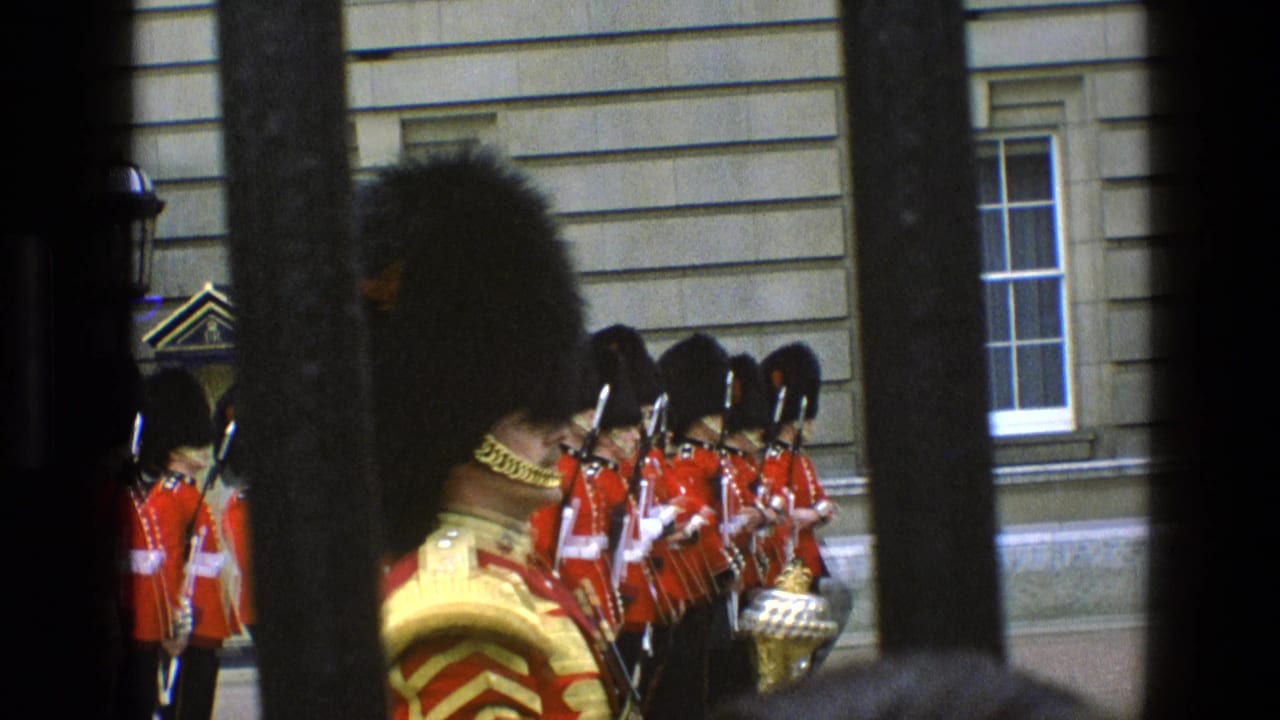 1969 London home-movie frame showing Queen's Guards in formation outside Buckingham Palace, partly framed by dark vertical bars.
