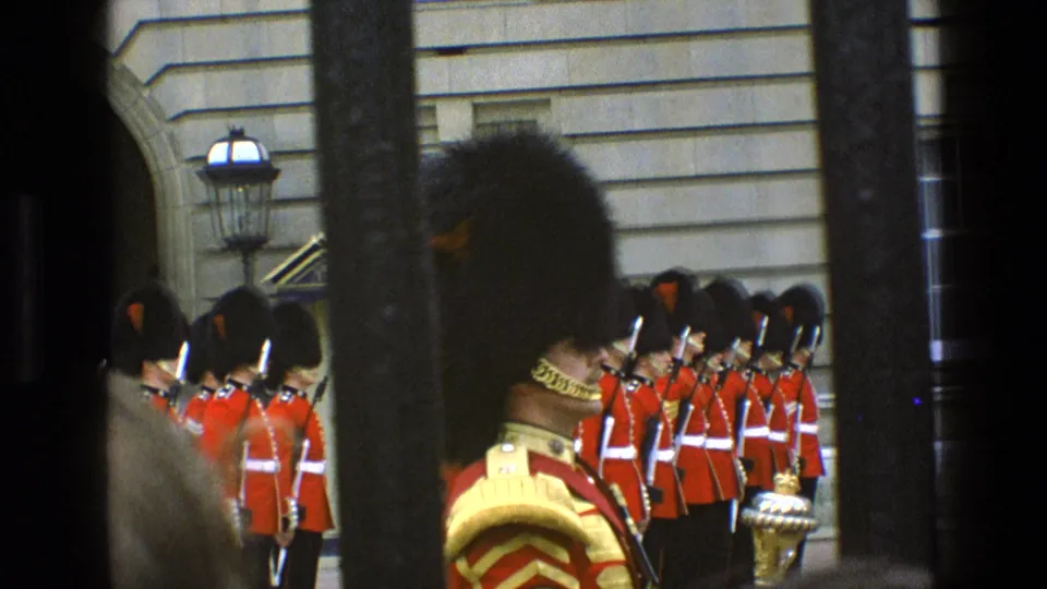 Obstructed 1969 London view of Queen's Guards outside Buckingham Palace, with a lamp and dark bars framing the formation.
