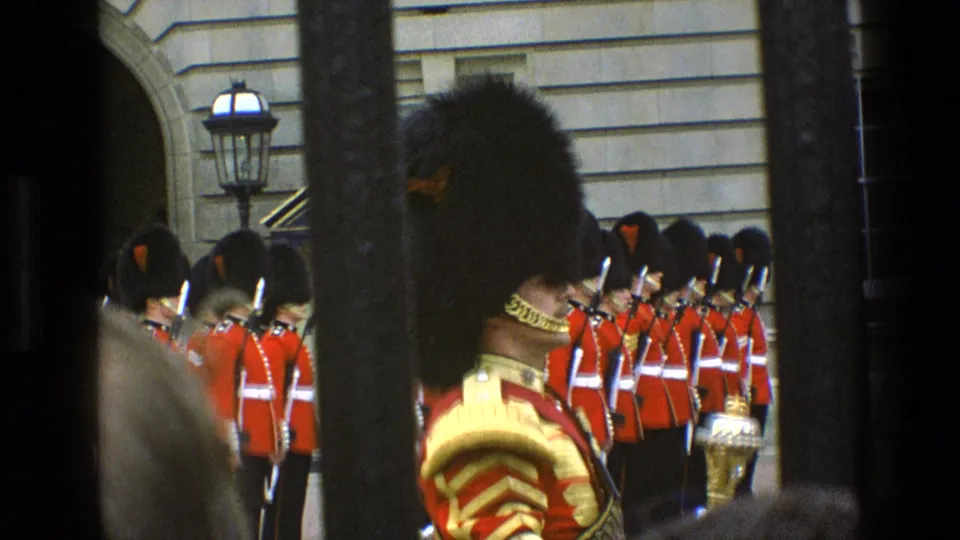 1969 home-movie frame of Buckingham Palace guards in red and gold ceremonial dress, seen through dark vertical railings.