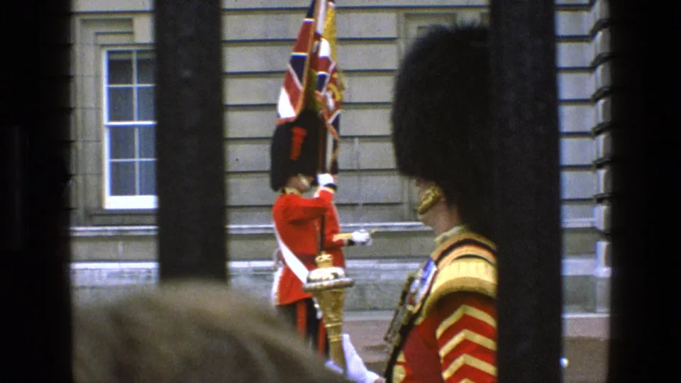 1969 England home-movie frame with a guard holding a Union Jack flag beside ceremonial uniformed figures and foreground bars.