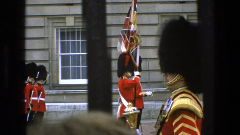 Ceremonial British guard with Union Jack flag appears near other red-coated guards, framed by dark bars in 1969 England footage.