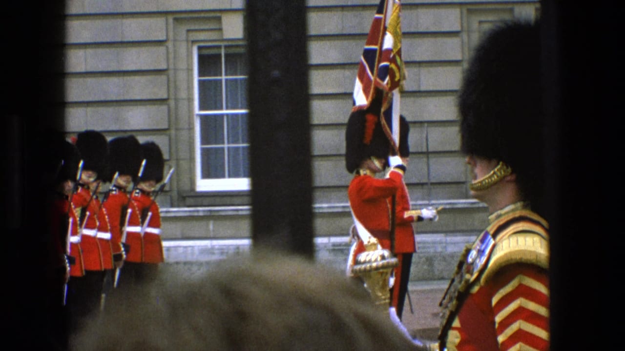 1969 England home-movie frame showing red-uniformed guards, bearskin hats, a Union Jack flag, and palace stonework seen through dark railings.