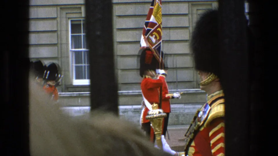 1969 England frame of the Changing of the Guard with red tunics, bearskin hats, ceremonial flag, and spectators partly obscuring the view.