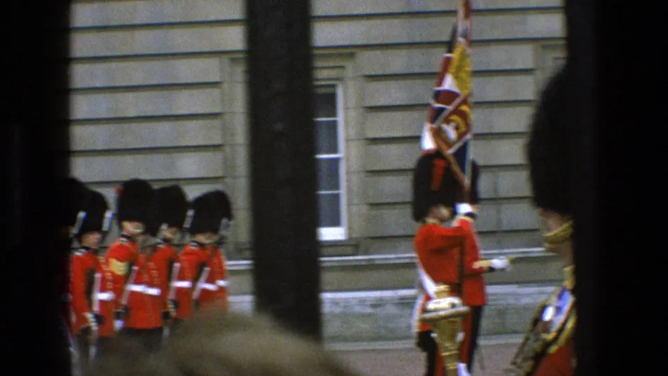 1969 England home-movie view of ceremonial guards in red uniforms, with a flag bearer and palace stonework partly obscured by railings.