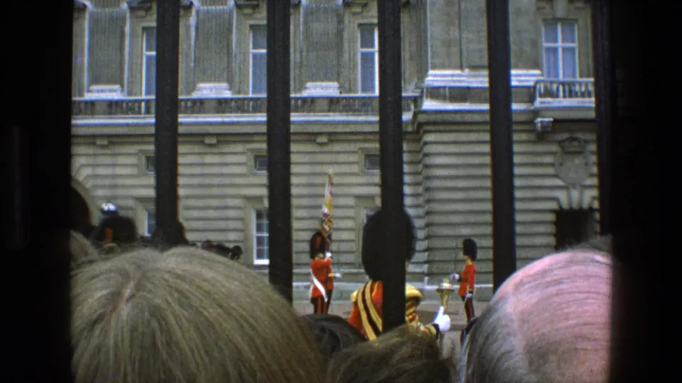 Wide 1969 England frame showing spectators behind railings watching red-coated guards and a flag bearer outside a grand stone building.